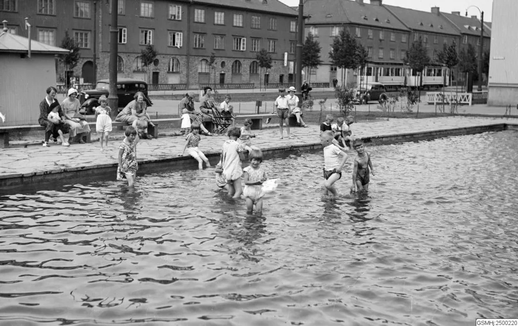 Svartvitt foto från 1935. Barn i sommarkläder vadar i den grunda plaskdammen på Älvsborgsplan. På kanten sitter mödrar med hattar, och bakom dem syns landshövdingehusens långa fasader, unga trädrader, en spårvagn och en bil.